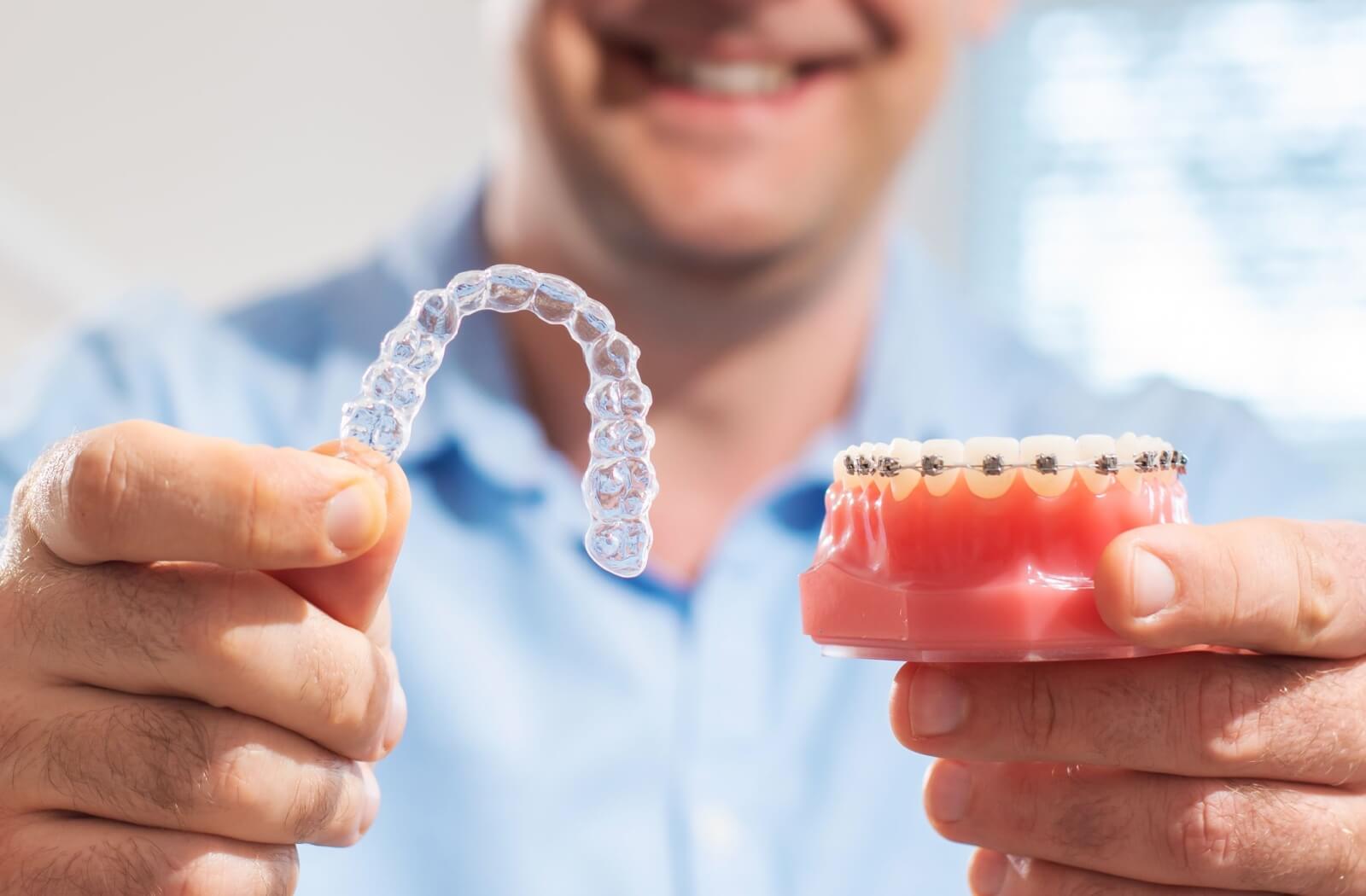 A smiling orthodontist in a blue shirt holds up an Invisalign mold and braces model to showcase crooked teeth treatments.